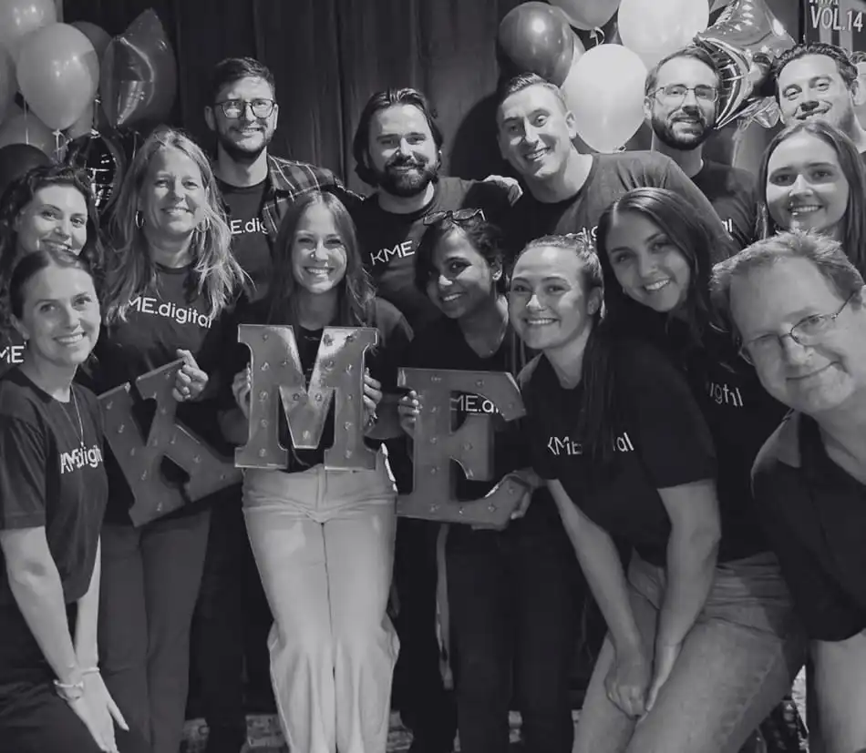 Team of coworkers posing at a celebration, holding large decorative letters 'M' and 'E' with balloons in the background.